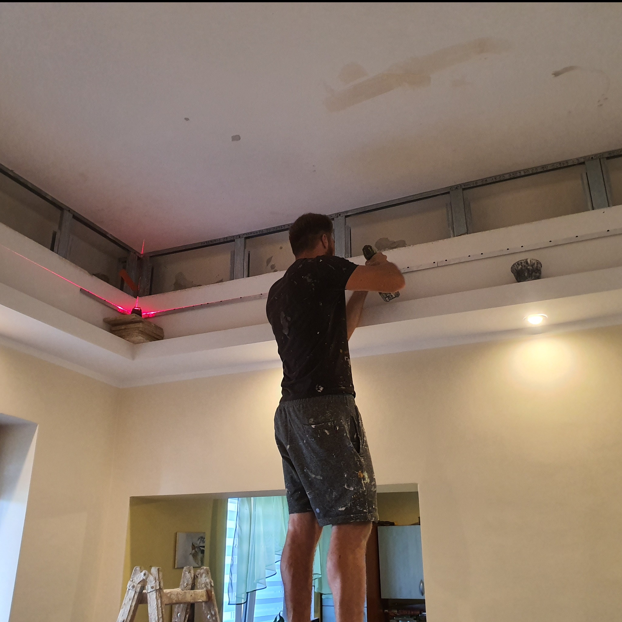 Pracownik na drabinie montuje konstrukcję sufitu podwieszanego, widoczny laser wyznaczający poziom, tło stanowią ściany w jasnym kolorze.