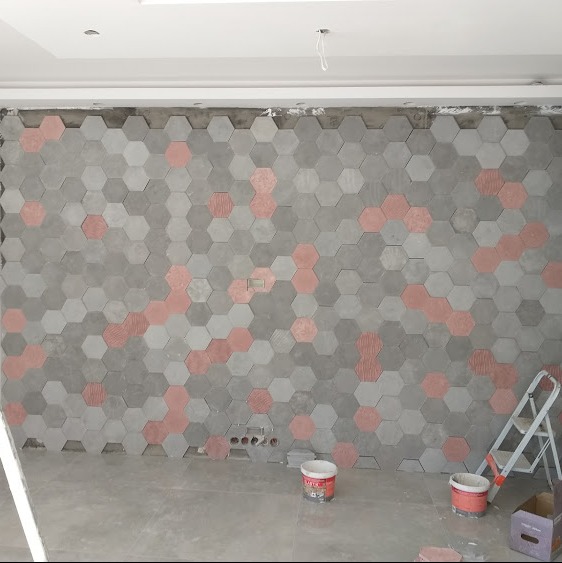 Ściana wyłożona szarymi i czerwonymi sześciokątnymi płytkami, widoczne puszki z farbą i drabina, instalacja w trakcie, widoczne otwory na gniazdka elektryczne.