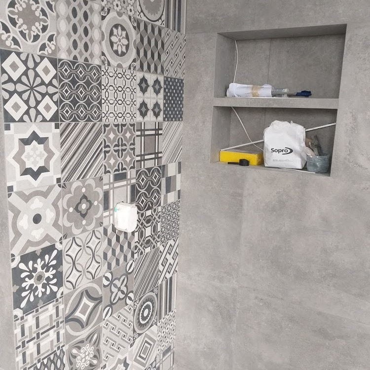 Ściana wyłożona wzorzystymi, szarymi płytkami ceramicznymi, obok wnęka z półkami, na których leżą białe ręczniki, żółta gąbka i worek z zaprawą Sopro.