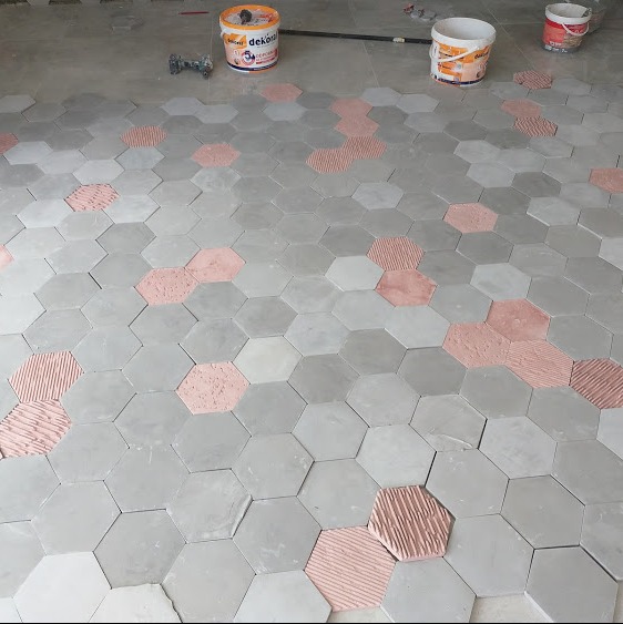 Układanie szarych, heksagonalnych płytek podłogowych z akcentami różowych płytek z nałożonym klejem, widoczne narzędzia i wiaderka z zaprawą.