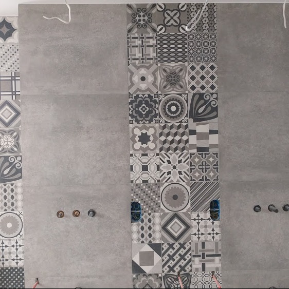 Szara ściana w trakcie wykańczania, z pasem dekoracyjnych, wzorzystych płytek ułożonych pionowo, widoczne wystające kable i rury, przygotowane otwory na armaturę.