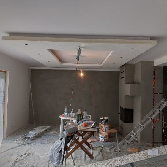 Pokój w trakcie remontu: surowe ściany, częściowo wykończony sufit podwieszany z oświetleniem, drabiny i narzędzia malarskie porozrzucane na podłodze zabezpieczonej folią.