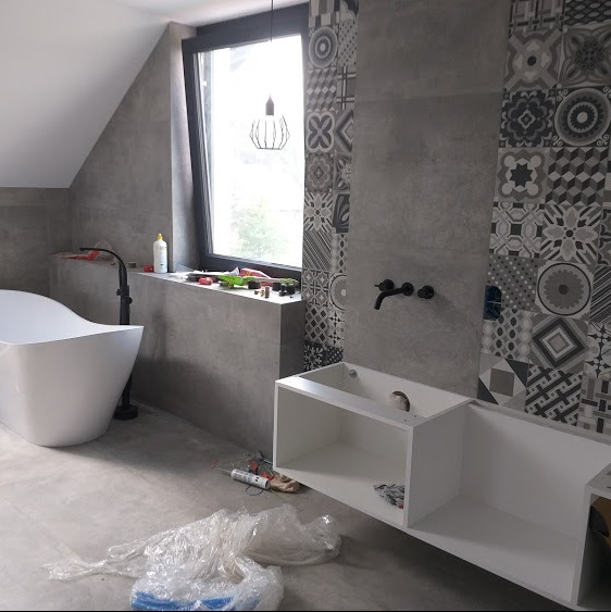 Nowoczesna łazienka w trakcie remontu: wolnostojąca wanna, szara ściana z wzorzystymi płytkami, biała zabudowa umywalki bez blatu, czarna bateria ścienna, narzędzia na parapecie okna.