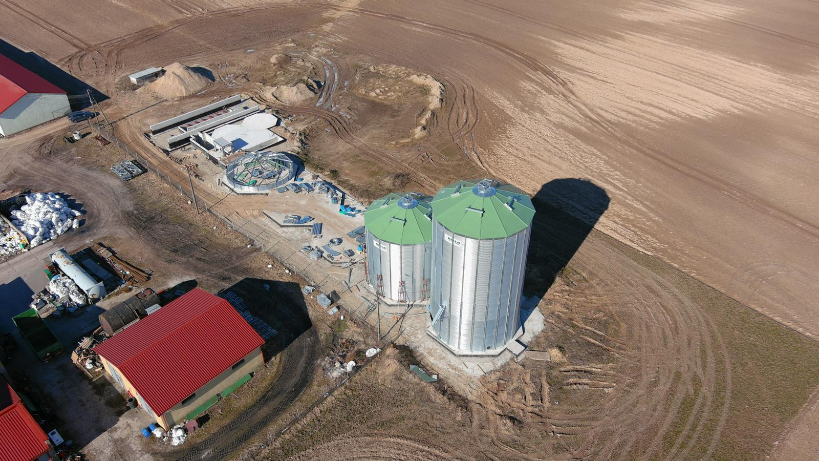 Widok z lotu ptaka na plac budowy w Żelistrzewie, gdzie widać dwa srebrne silosy z zielonymi dachami oraz fundamenty pod inne konstrukcje. Na placu widoczne są materiały budowlane i ślady opon...