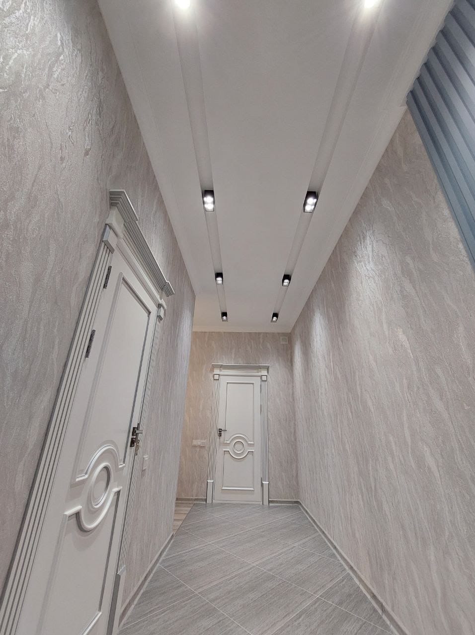 Korytarz po remoncie z szarymi ścianami o fakturze imitującej kamień, białe drzwi z dekoracyjnymi ramami i listwami, oraz podłoga wyłożona szarymi płytkami imitującymi drewno, oświetlenie punktowe...