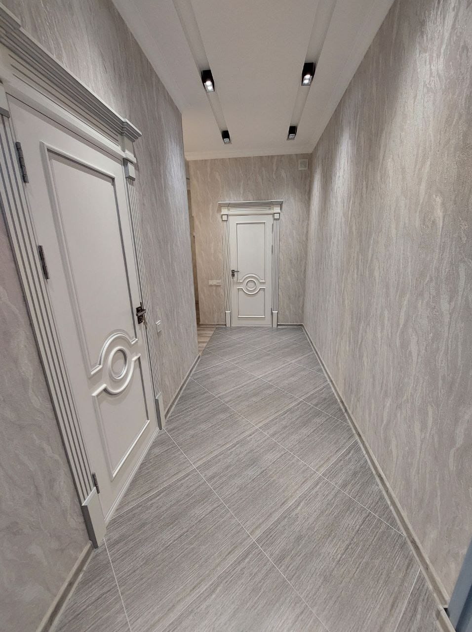 Korytarz po remoncie z szarymi płytkami na podłodze ułożonymi pod kątem, ściany pokryte tapetą o strukturze kamienia, białe drzwi z dekoracyjnymi ramami i czarne reflektory sufitowe.