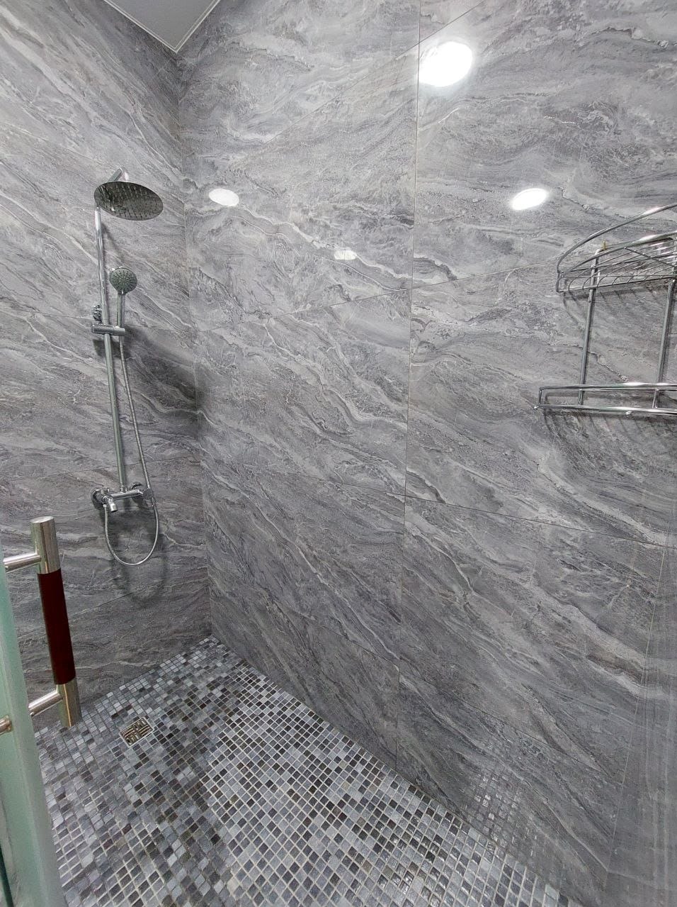 Nowoczesny prysznic z szarą marmurkową okładziną ścienną i mozaikową podłogą, chromowana armatura i półka narożna.
