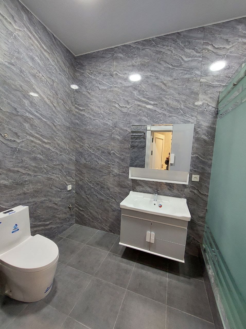 Szara łazienka z marmurowymi płytkami na ścianach, białą toaletą i szafką pod umywalkę z lustrem.