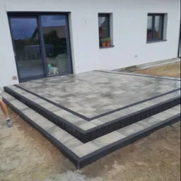 Nowoczesny taras z kostki brukowej w odcieniach szarości, z dwustopniowymi schodami, obrzeżami w kolorze antracytowym i widocznym fragmentem nogi osoby w lewym dolnym rogu.