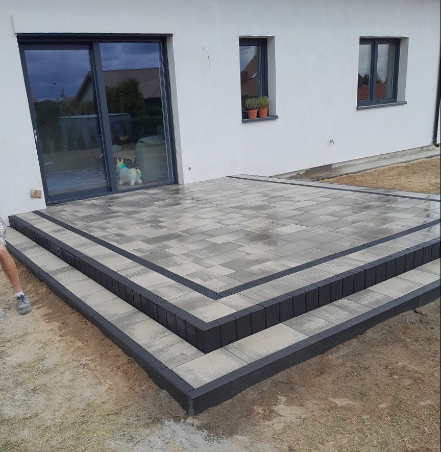 Nowoczesny taras z kostki brukowej w odcieniach szarości, z dwustopniowymi schodami, obrzeżami w kolorze antracytowym i widocznym fragmentem nogi osoby w lewym dolnym rogu.