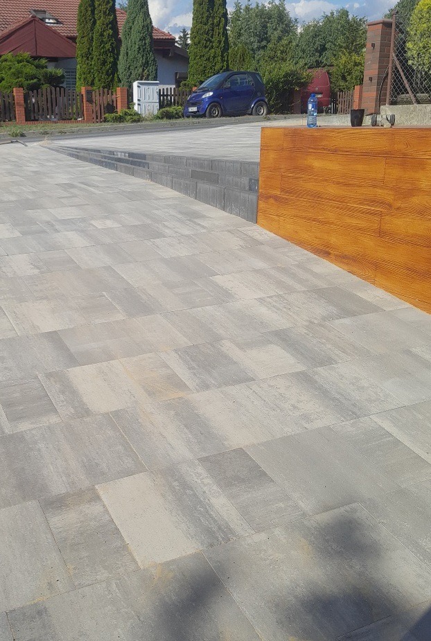 Nowoczesny podjazd z szarej kostki brukowej ułożonej w geometryczny wzór, z widocznym podjazdem do domu i zaparkowanym niebieskim samochodem w tle.