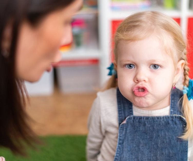 Dziewczynka w dżinsowej sukience ćwiczy wymowę z logopedą, skupiona twarz i ułożenie ust podczas artykulacji głoski.