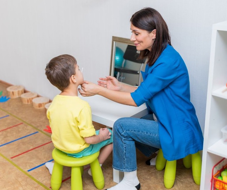 Chłopiec siedzi na zielonym krześle podczas badania jamy ustnej przez kobietę w niebieskiej bluzce, białe ściany i podłoga z kolorowymi liniami w tle.