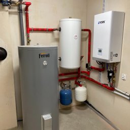 Notos Engenering Polska Sp z o.o. - Instalacja grzewcza w kotłowni: szary pionowy zbiornik Ferroli, biały zbiornik akumulacyjny i biały piec York połączone czerwonymi rurami, z małymi niebieskimi i białymi zbiornikami wyrównawczymi.