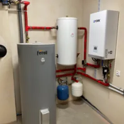 Instalacja grzewcza w kotłowni: szary pionowy zbiornik Ferroli, biały zbiornik akumulacyjny i biały piec York połączone czerwonymi rurami, z małymi niebieskimi i białymi zbiornikami wyrównawczymi.
