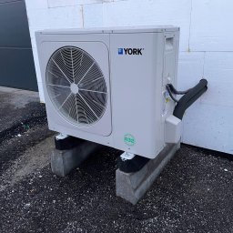 Notos Engenering Polska Sp z o.o. - Zewnętrzna jednostka pompy ciepła marki York, zamontowana na betonowych podstawach przy ścianie budynku z widocznymi rurami instalacji.