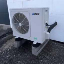 Zewnętrzna jednostka pompy ciepła marki York, zamontowana na betonowych podstawach przy ścianie budynku z widocznymi rurami instalacji.