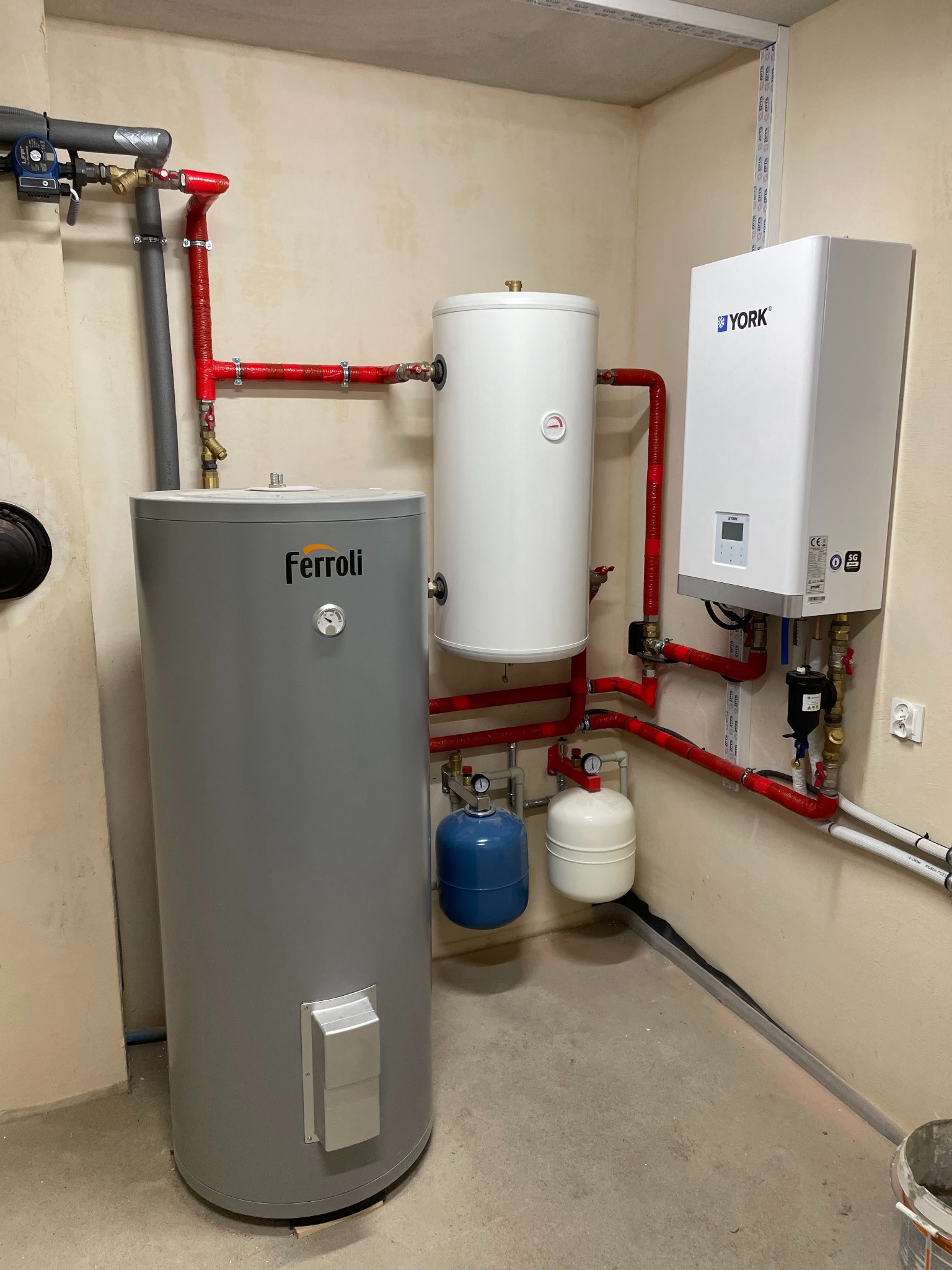 Instalacja grzewcza w kotłowni: szary pionowy zbiornik Ferroli, biały zbiornik akumulacyjny i biały piec York połączone czerwonymi rurami, z małymi niebieskimi i białymi zbiornikami wyrównawczymi.