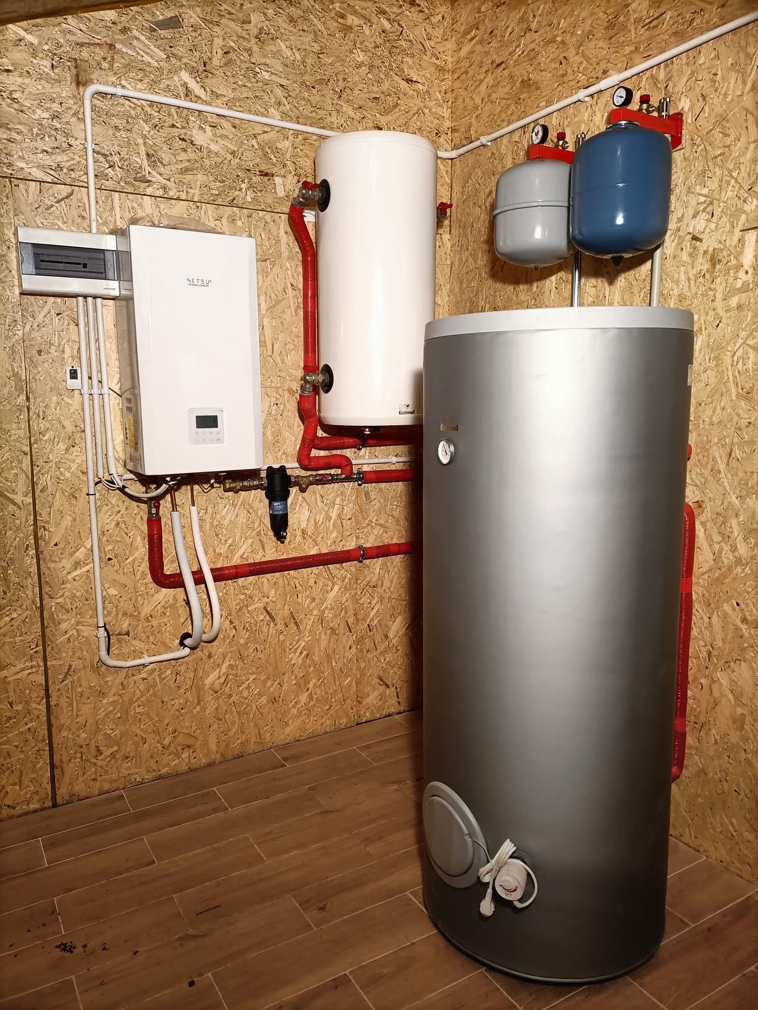 Kotłownia z zamontowanym zasobnikiem ciepłej wody, kotłem grzewczym oraz naczyniami przeponowymi, widoczne rury w kolorze czerwonym i białym, ściany wykończone płytami OSB.
