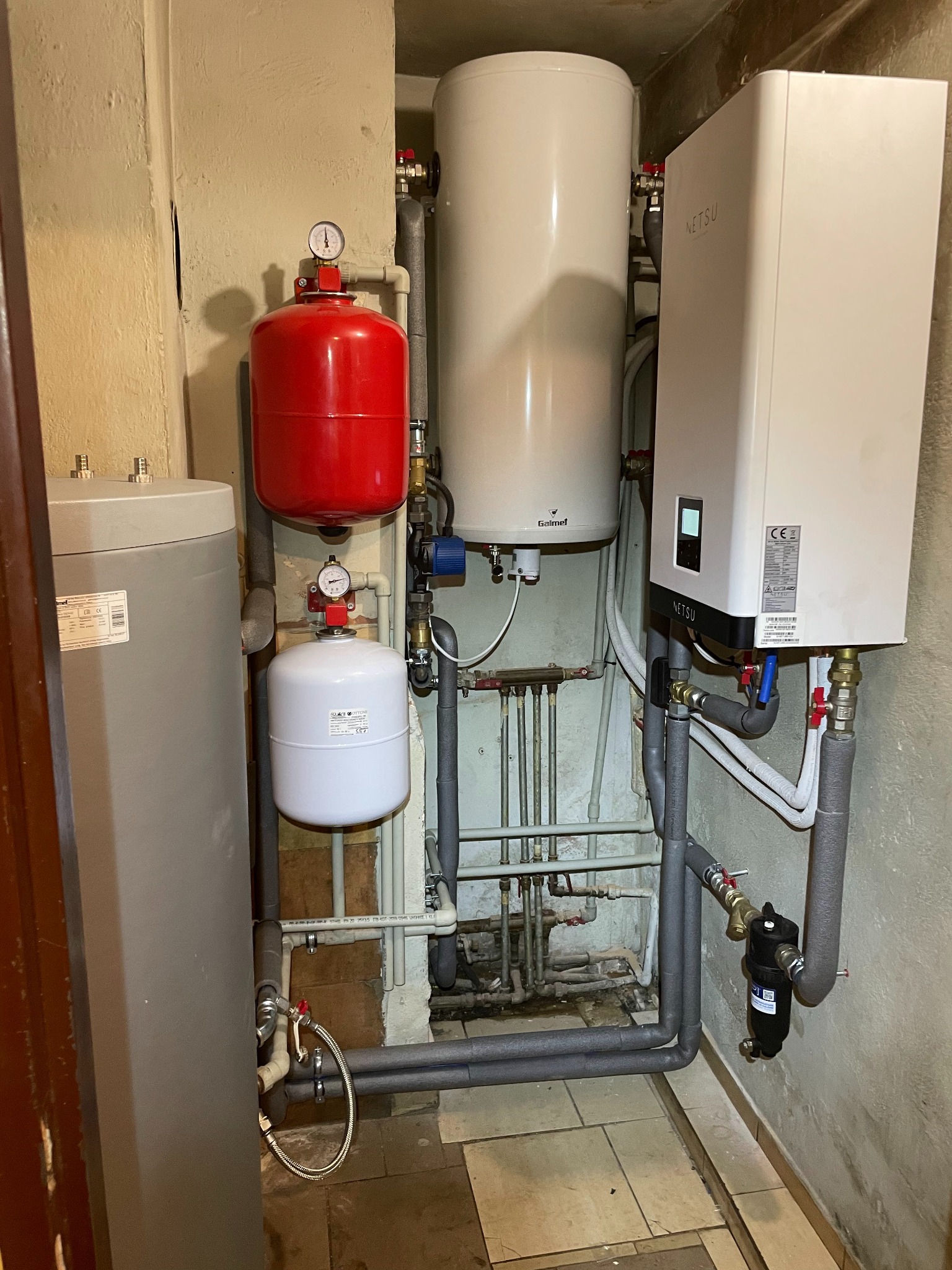 Instalacja grzewcza w piwnicy: biały zasobnik ciepłej wody Galmet, czerwony i biały zbiornik przeponowy Reflex z manometrami, szary bojler, biała jednostka NETSU z wyświetlaczem, rury w szarej...