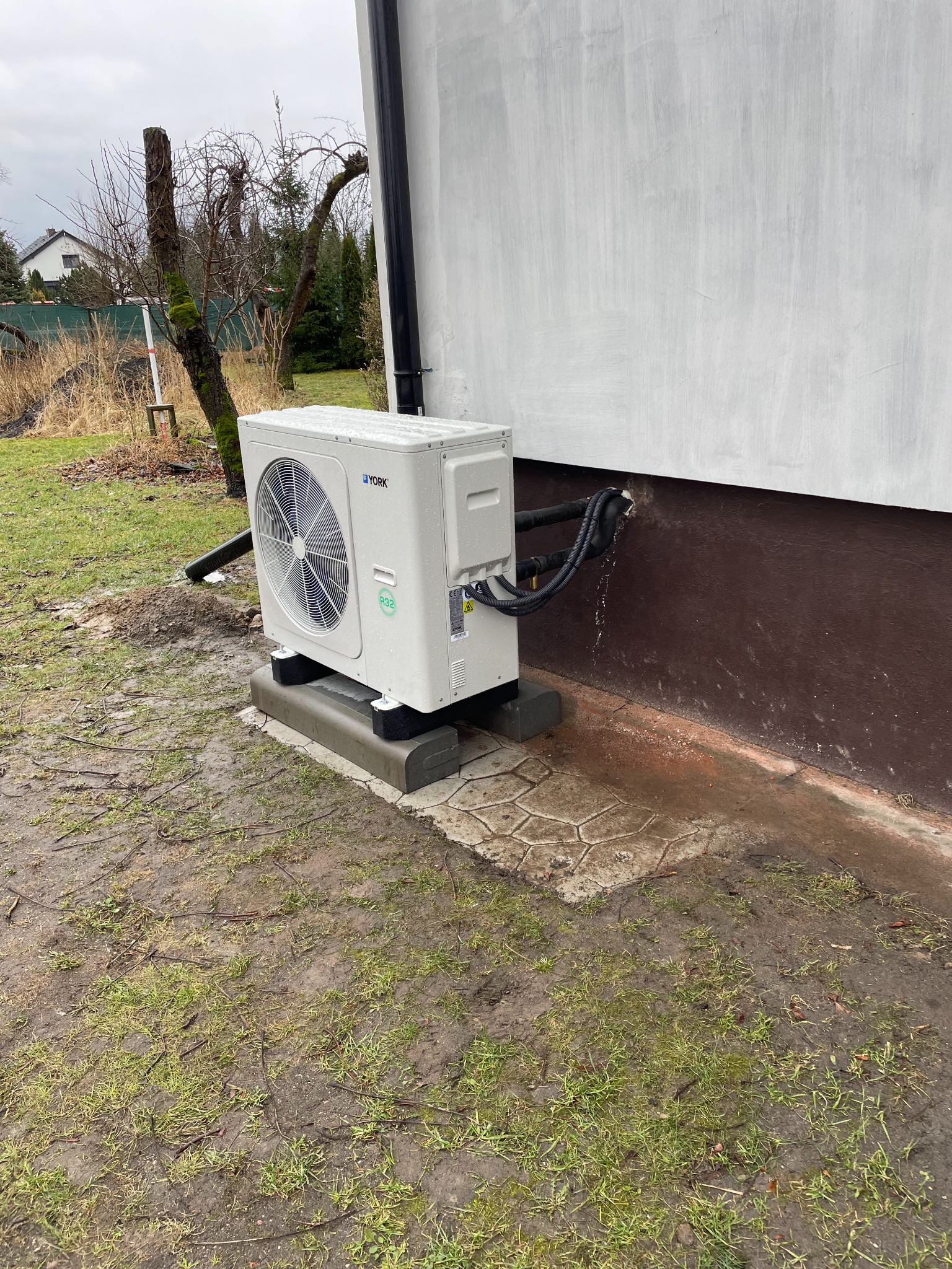 Biała jednostka zewnętrzna pompy ciepła marki York, zamontowana na betonowej podstawie przy ścianie budynku z widocznymi przyłączami rur. W tle trawnik z błotem i drzewo bez liści.
