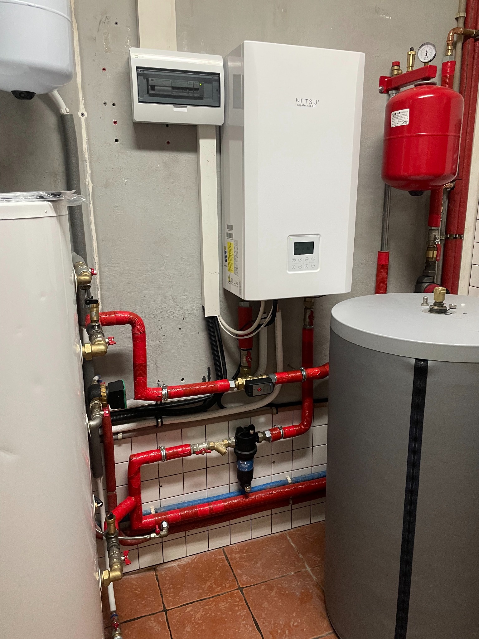 Biała pompa ciepła marki Netsu zamontowana na szarej ścianie, z widocznymi czerwonymi rurami instalacji hydraulicznej, zbiornikiem wyrównawczym i bojlerem, całość na tle terakoty.
