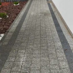 Szara kostka brukowa z ciemniejszymi pasami po bokach, ułożona wzdłuż białej ściany budynku i rabaty z korą drzewną, widok z góry.