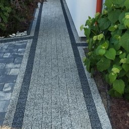 PS Projekt Mycie ciśnieniowe - Ułożona ścieżka z kostki brukowej w odcieniach szarości, obramowana ciemniejszymi pasami, prowadząca wzdłuż ściany budynku z kwiatami w oknie i zielenią po bokach.