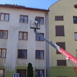 Pracownik w kombinezonie ochronnym na podnośniku koszowym czyści elewację budynku, widoczne różnice w kolorze między umytą a brudną częścią ściany.
