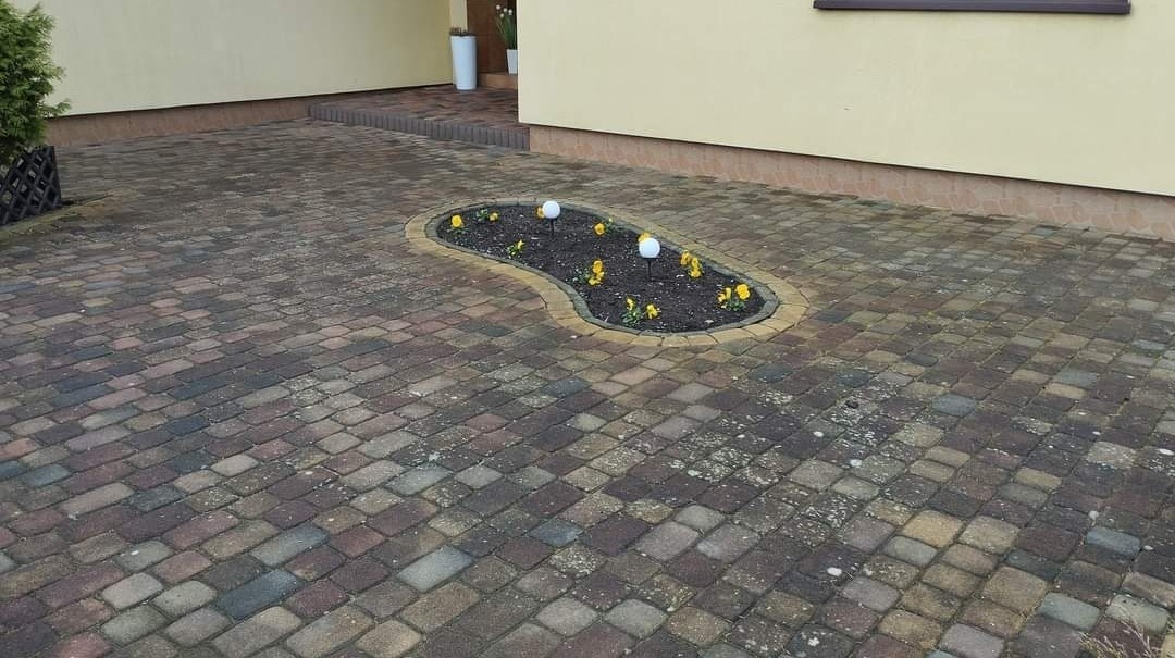 Wielobarwna kostka brukowa prowadząca do wejścia domu, z małym klombem w kształcie nerki obsadzonym żółtymi kwiatami i oświetlonym dwoma białymi lampami solarnymi.