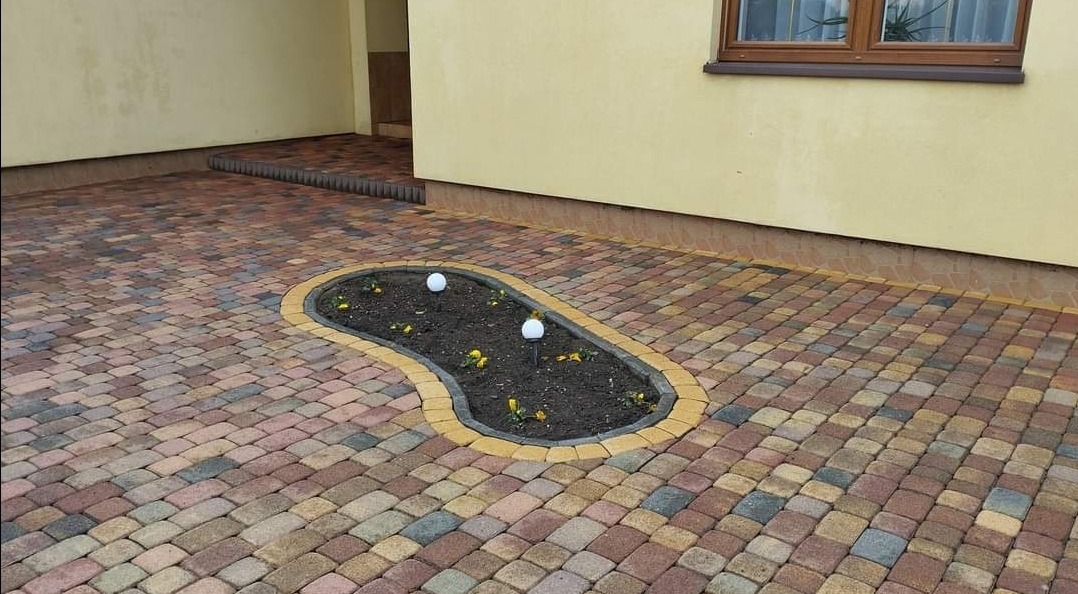 Kolorowa kostka brukowa ułożona na podjeździe z wyznaczoną rabatą kwiatową w kształcie nerki, oświetlona dwoma lampami solarnymi.
