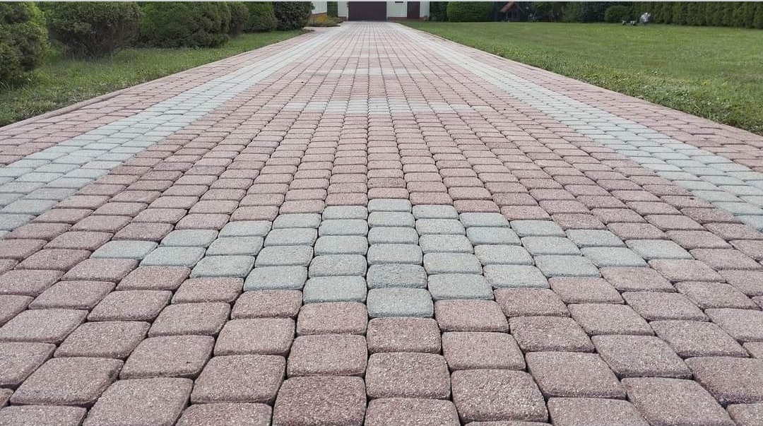 Ułożona z kostki brukowej aleja w odcieniach czerwieni i szarości, prowadząca w kierunku domu z ciemnymi drzwiami garażowymi, otoczona zielonym trawnikiem i krzewami.