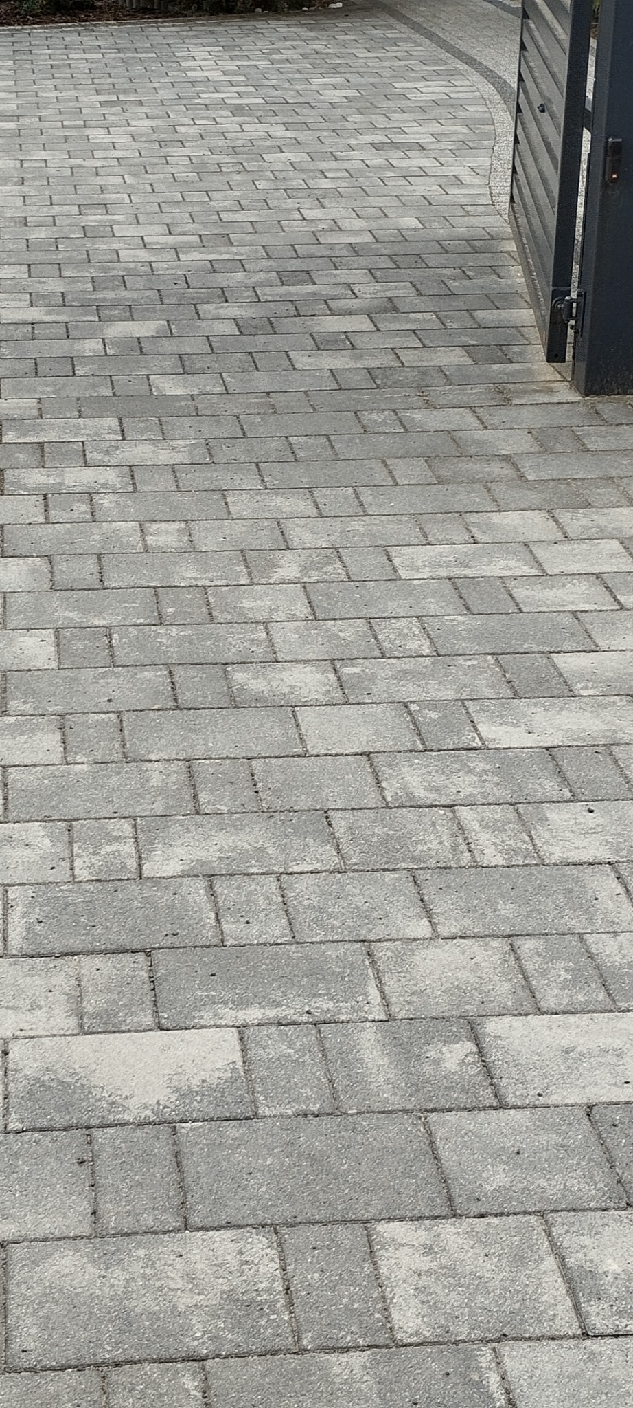 Szara kostka brukowa ułożona w regularny wzór w Bełchatowie, widoczna brama w tle, częściowo wyczyszczona powierzchnia kostki.