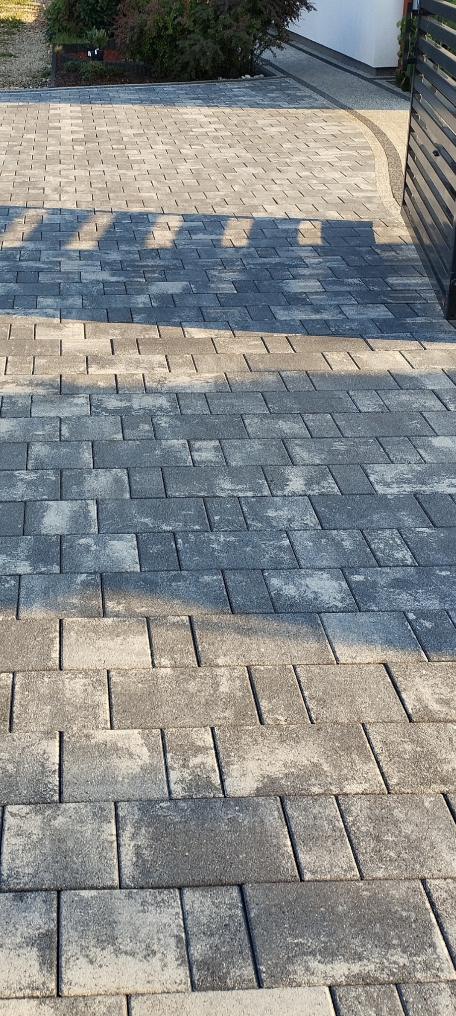 Ułożona szara kostka brukowa na podjeździe, częściowo zacieniona, z widoczną bramą w tle.