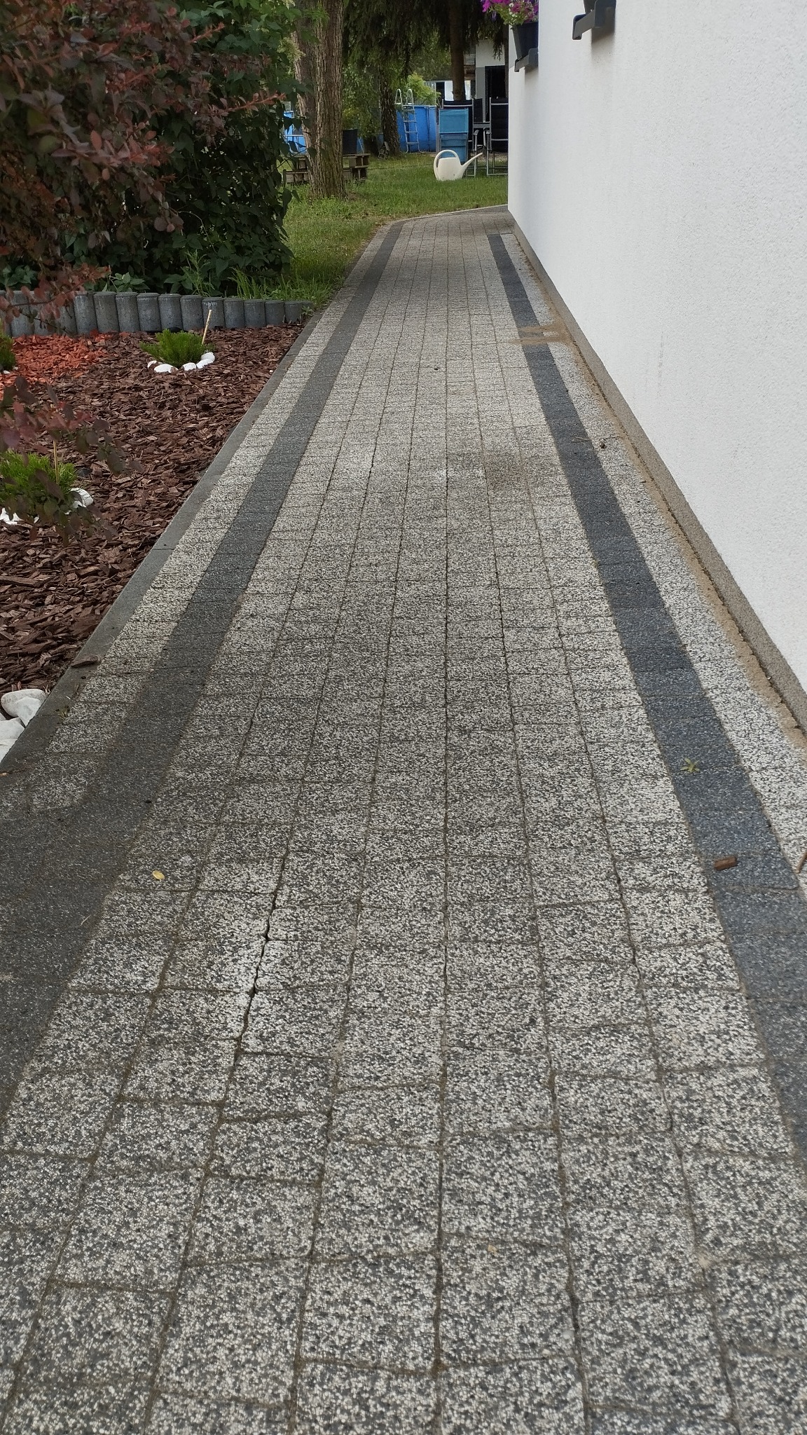 Szara kostka brukowa z ciemniejszymi pasami po bokach, ułożona wzdłuż białej ściany budynku i rabaty z korą drzewną, widok z góry.
