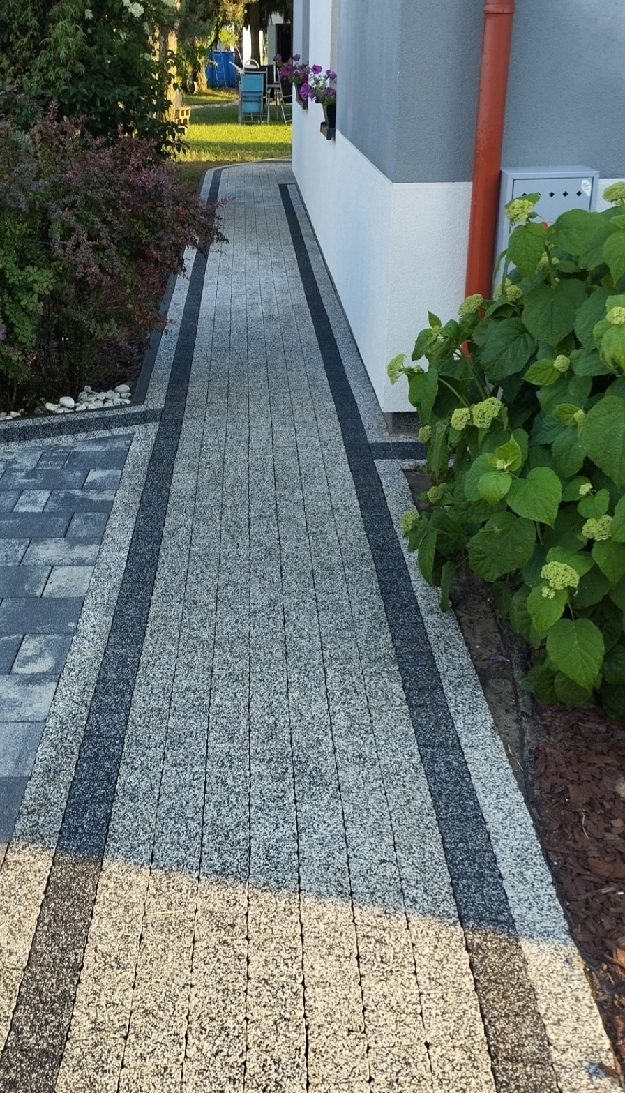Ułożona ścieżka z kostki brukowej w odcieniach szarości, obramowana ciemniejszymi pasami, prowadząca wzdłuż ściany budynku z kwiatami w oknie i zielenią po bokach.