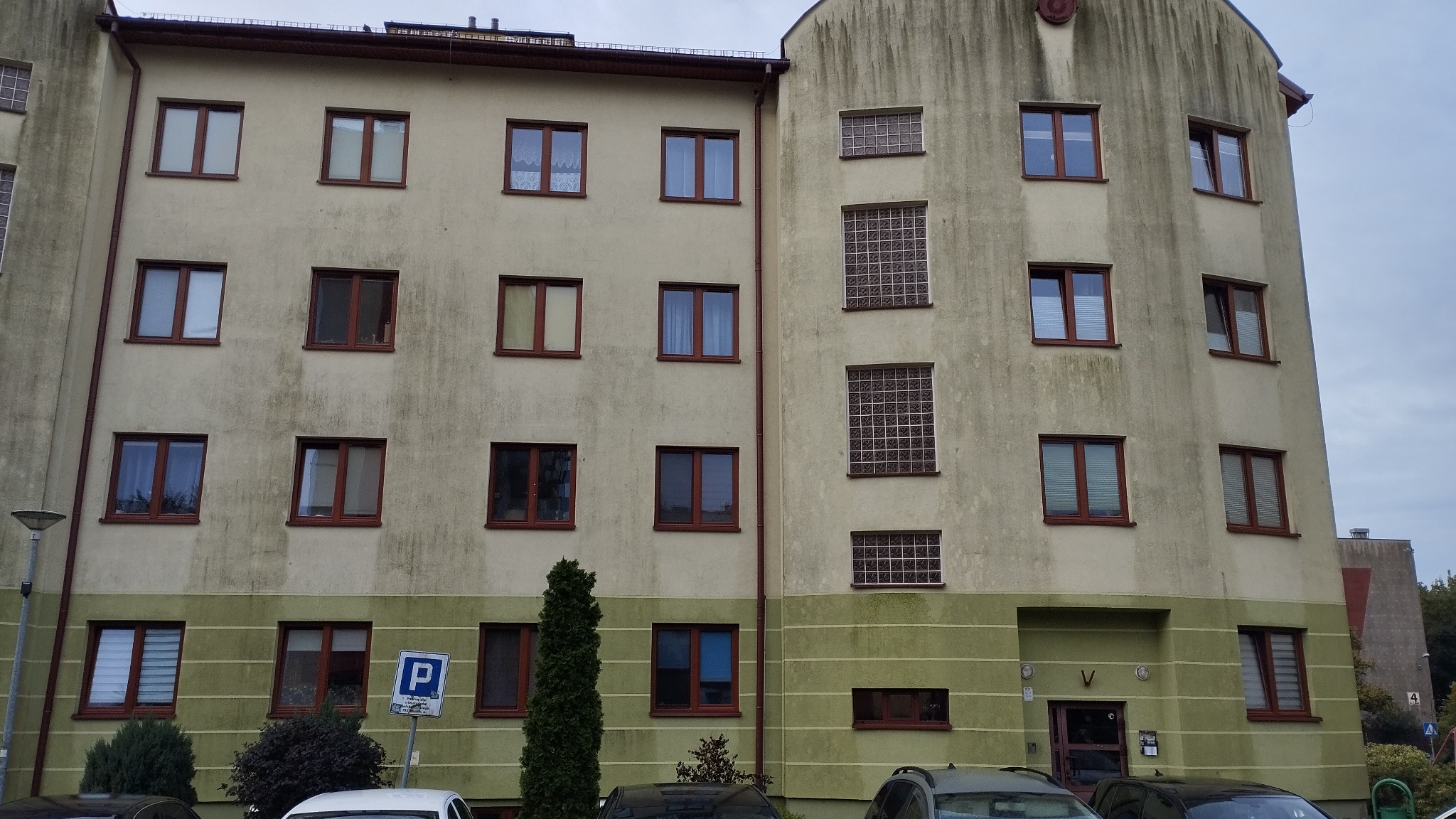 Wielopiętrowy budynek mieszkalny z widocznymi śladami zabrudzeń na elewacji, zwłaszcza w postaci zacieków i ciemnych plam, z oknami o brązowych ramach i częściowo zasłoniętymi roletami,...