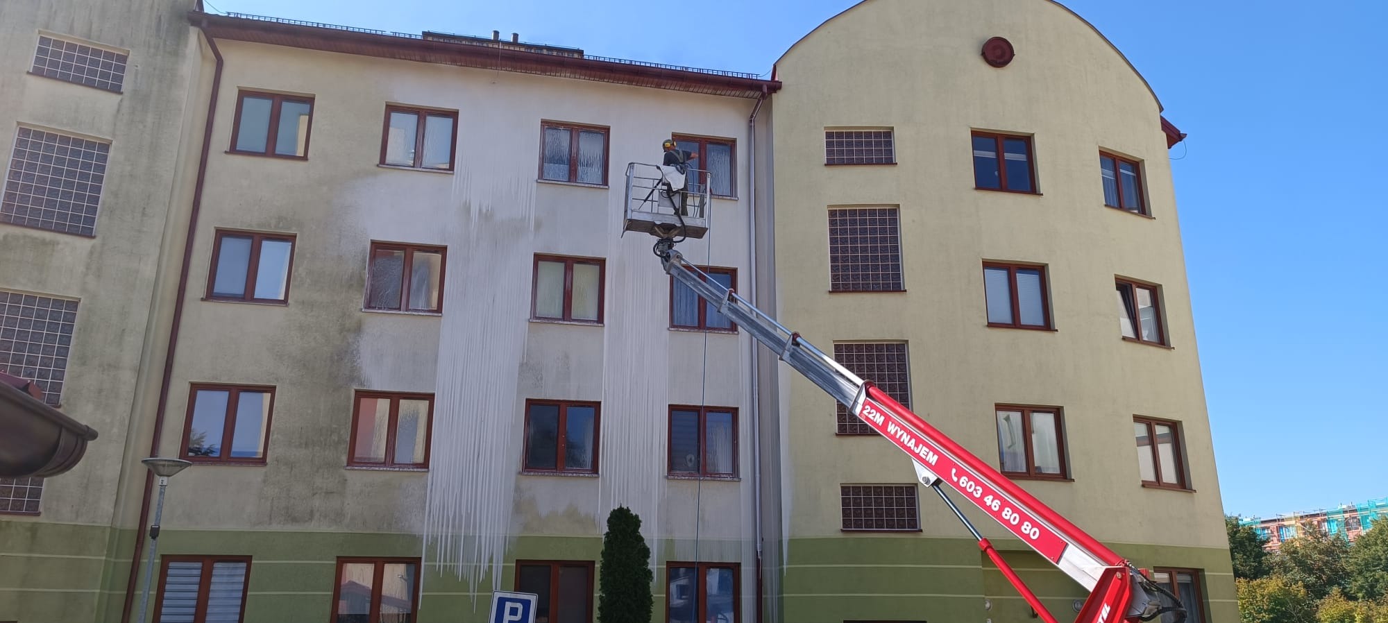 Pracownik w kombinezonie ochronnym na podnośniku koszowym czyści elewację budynku, widoczne różnice w kolorze między umytą a brudną częścią ściany.