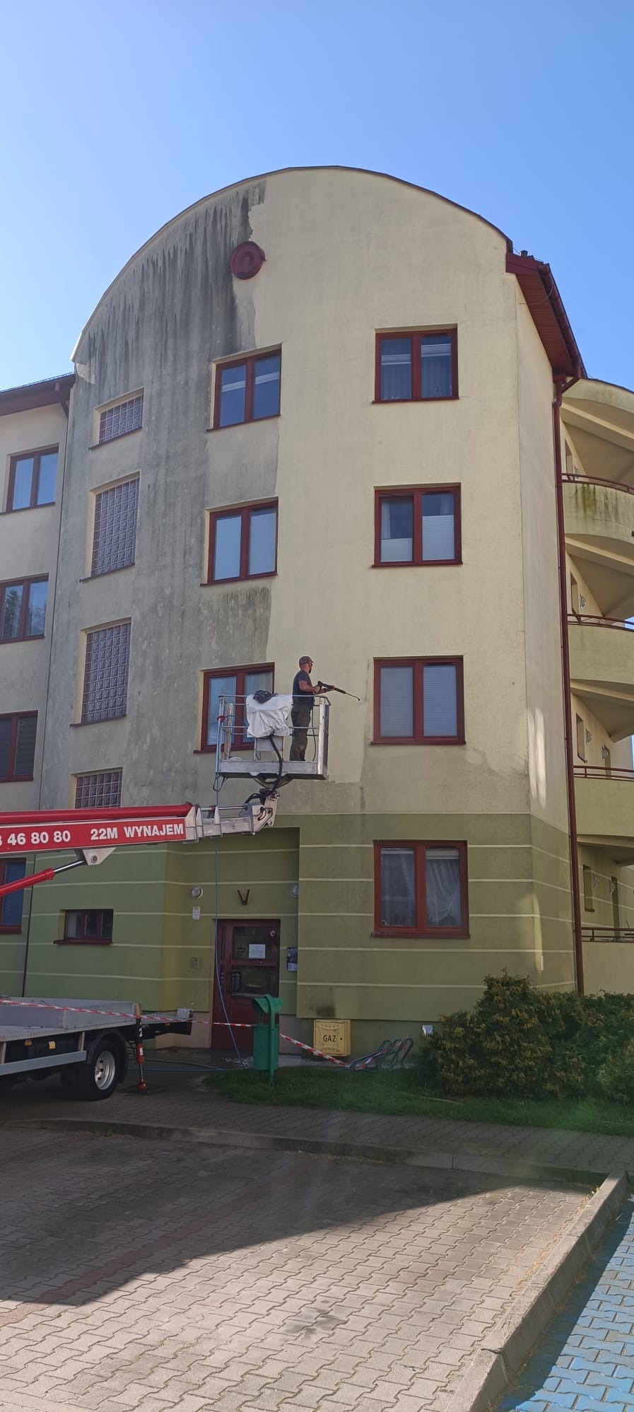 Pracownik na podnośniku koszowym czyści elewację bloku mieszkalnego za pomocą myjki ciśnieniowej, widoczne różnice w kolorze przed i po czyszczeniu.