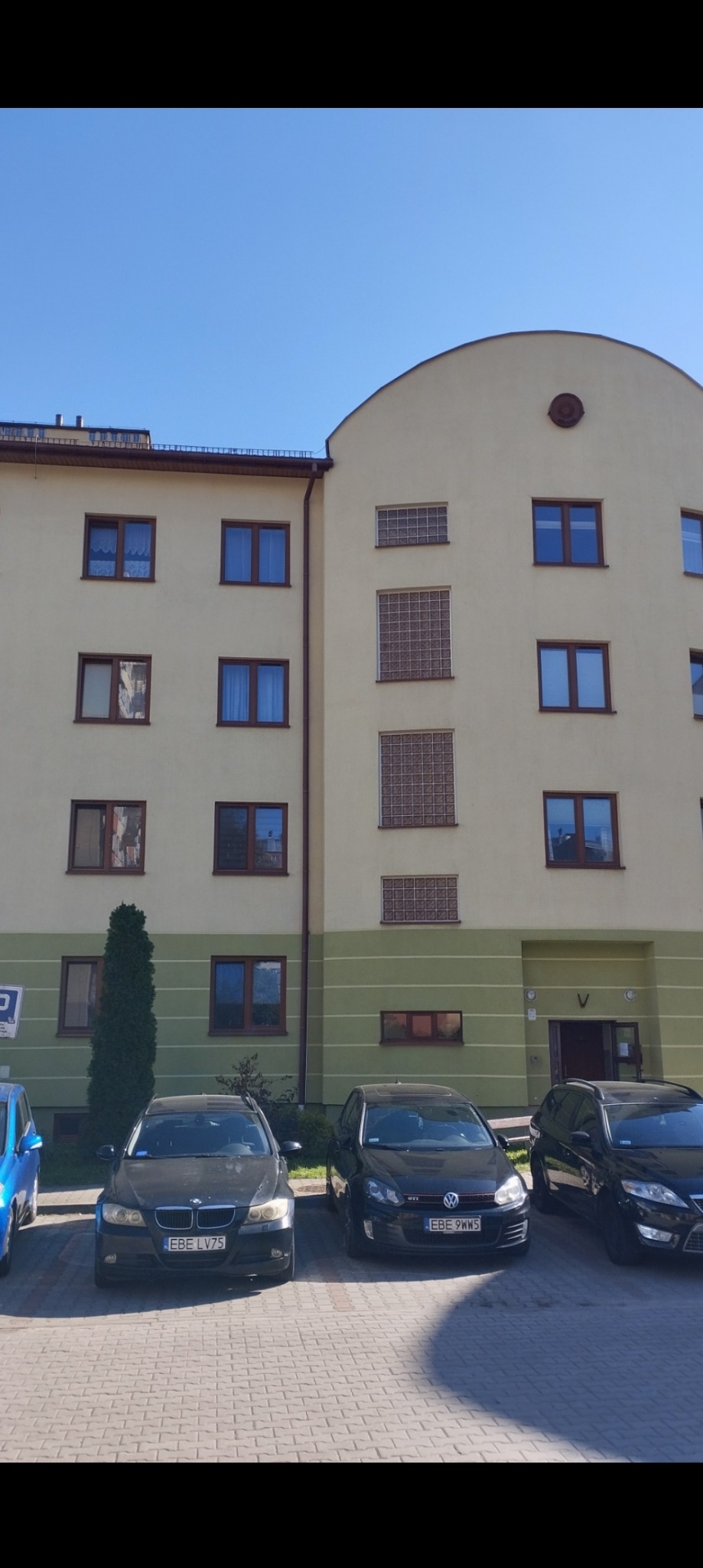 Budynek mieszkalny o żółtej elewacji z zielonym pasem na dole, z widocznymi oknami i trzema zaparkowanymi samochodami na kostce brukowej.