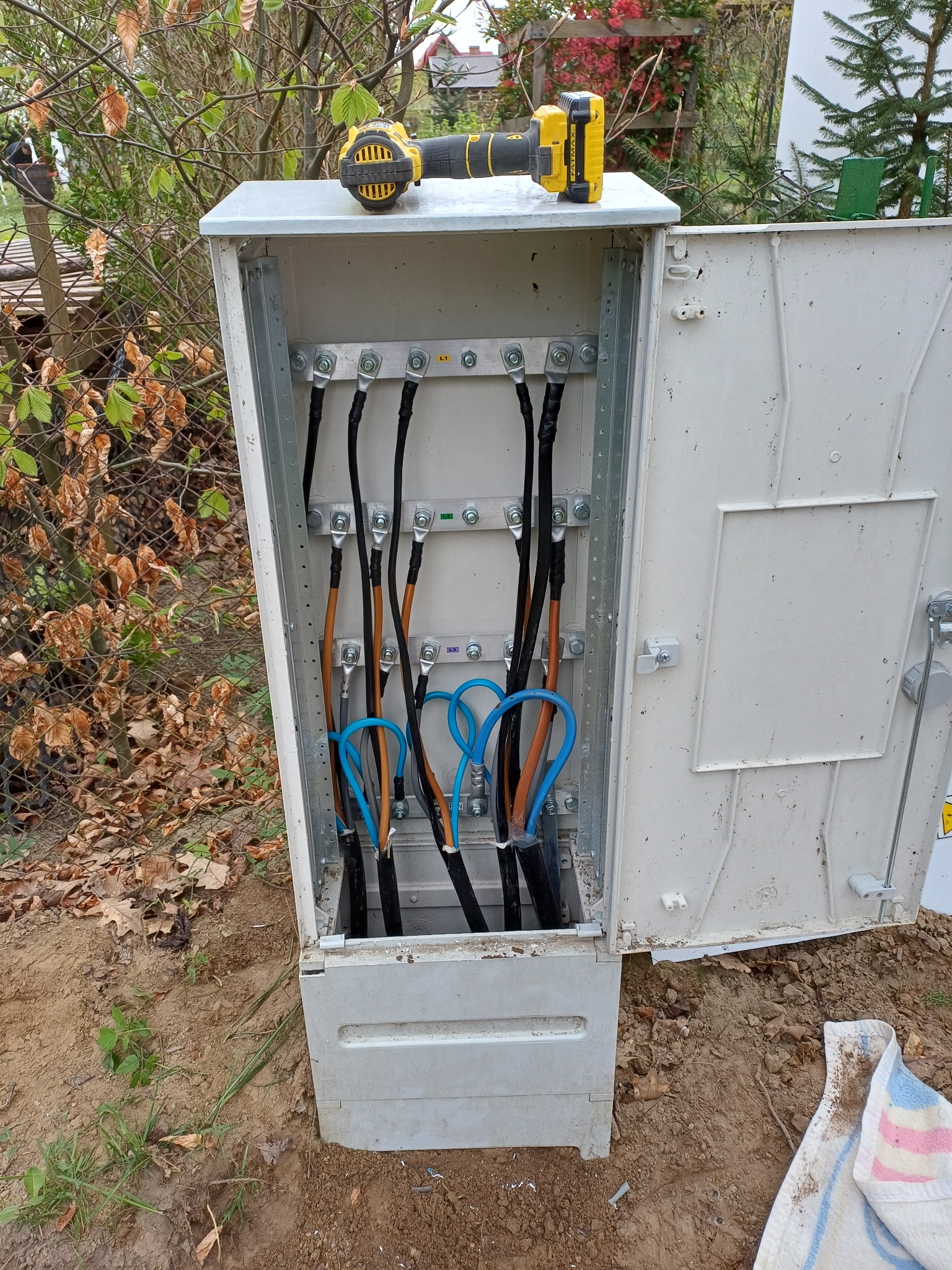Otwarta skrzynka elektryczna z podłączonymi przewodami, na górze leży wkrętarka akumulatorowa, widok na zewnątrz w tle.