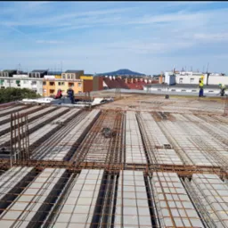 Prace na budowie: zbrojenie stropu Teriva z widocznymi pracownikami w żółtych kamizelkach i kaskach na tle miejskiego krajobrazu z budynkami i wzgórzem.