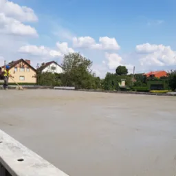 Świeżo wylany, wyrównywany fundament pod budowę domu jednorodzinnego, pracownik w kamizelce odblaskowej i kasku, na tle zabudowy mieszkalnej.