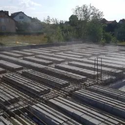 Budowa stropu Teriva: pracownik w kasku i kamizelce odblaskowej przechodzi po konstrukcji z belek i pustaków, widoczne zbrojenie, w tle budynki mieszkalne i zieleń.