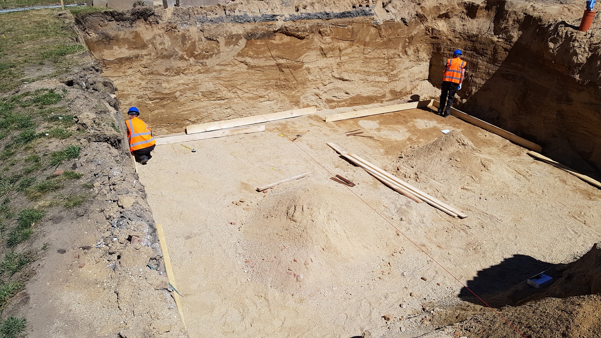 Przygotowanie wykopu pod fundamenty: Dwóch robotników w kamizelkach i kaskach, wytyczanie obrysu, drewniane deski szalunkowe w wykopie, widoczne warstwy gruntu.