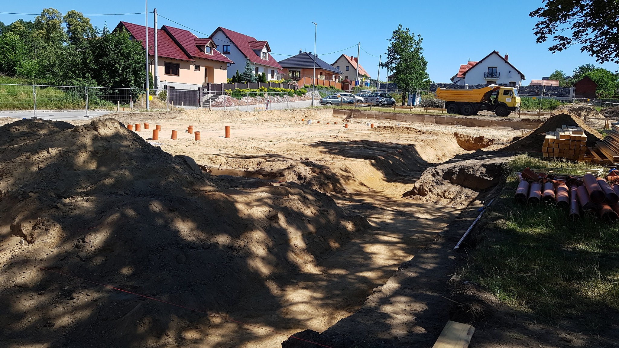 Wykonany wykop pod fundamenty domu jednorodzinnego, widoczne rury kanalizacyjne, hałda ziemi oraz żółta wywrotka w tle.