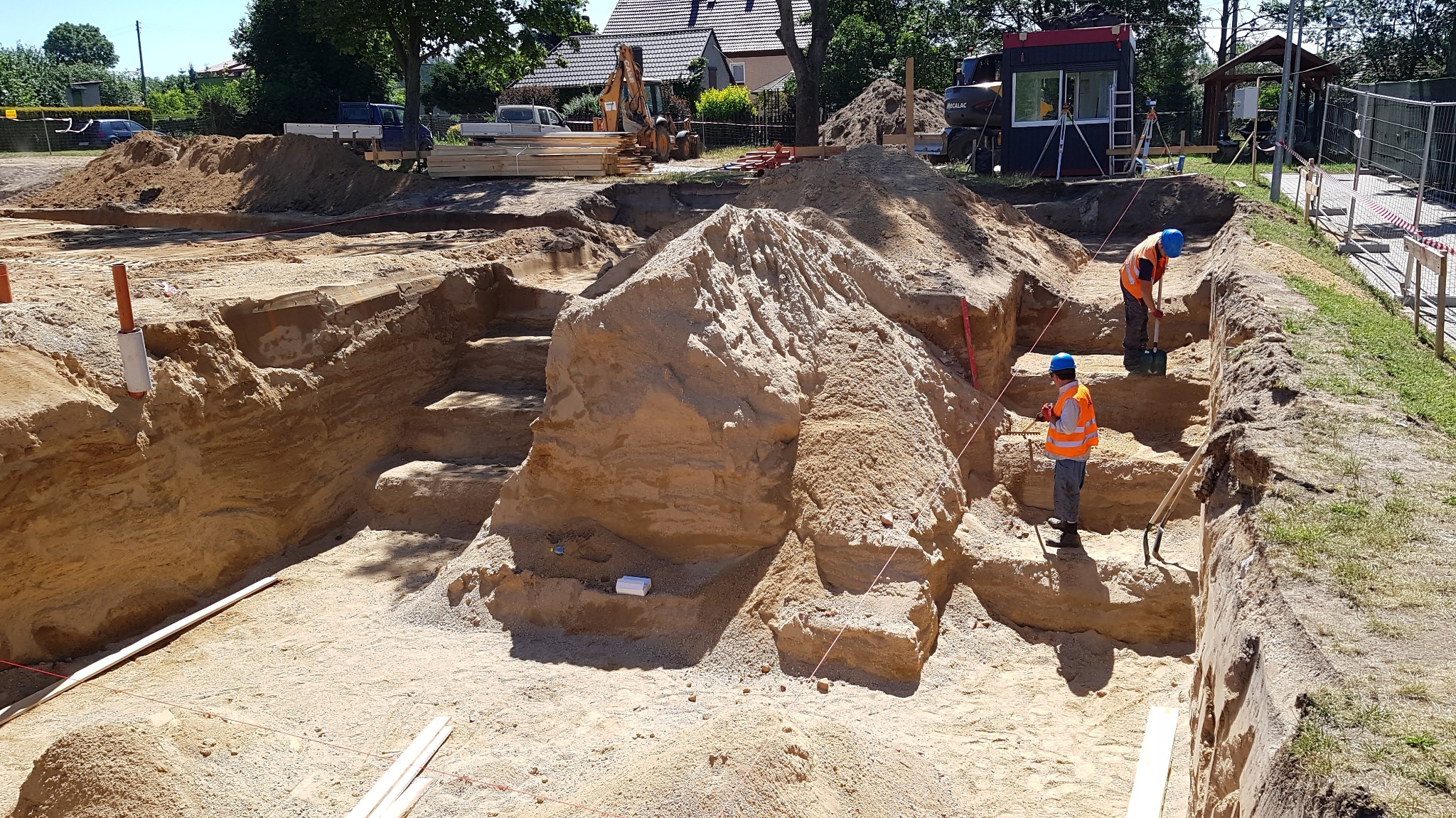 Wykop pod fundamenty z widocznymi warstwami gruntu, dwoma pracownikami w kaskach i kamizelkach odblaskowych podczas prac ziemnych, z użyciem szpadla i niwelatora, oraz sprzętem budowlanym w tle.