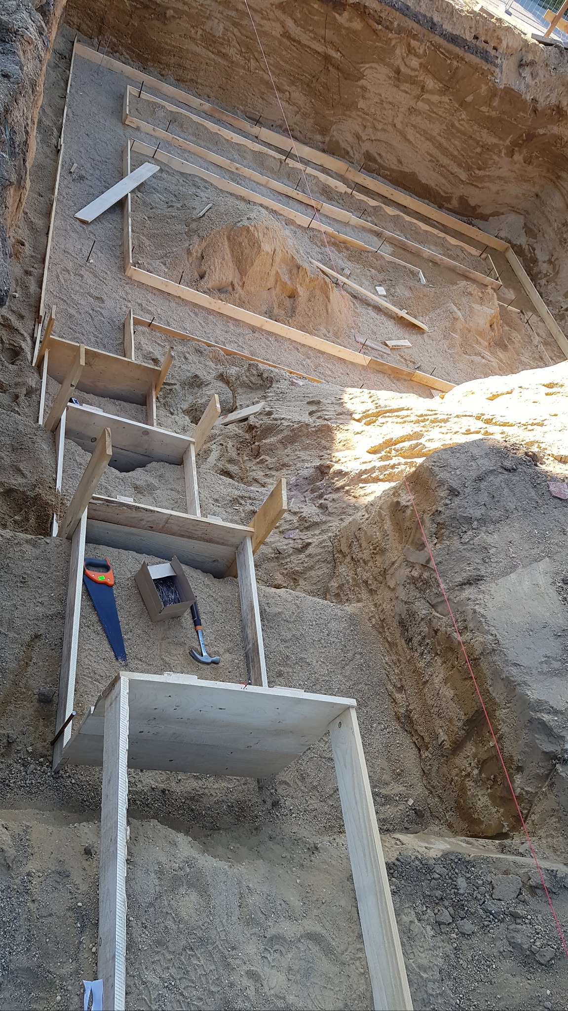 Wykop pod fundamenty z drewnianym szalunkiem, widoczna piła ręczna, młotek i pudełko gwoździ.