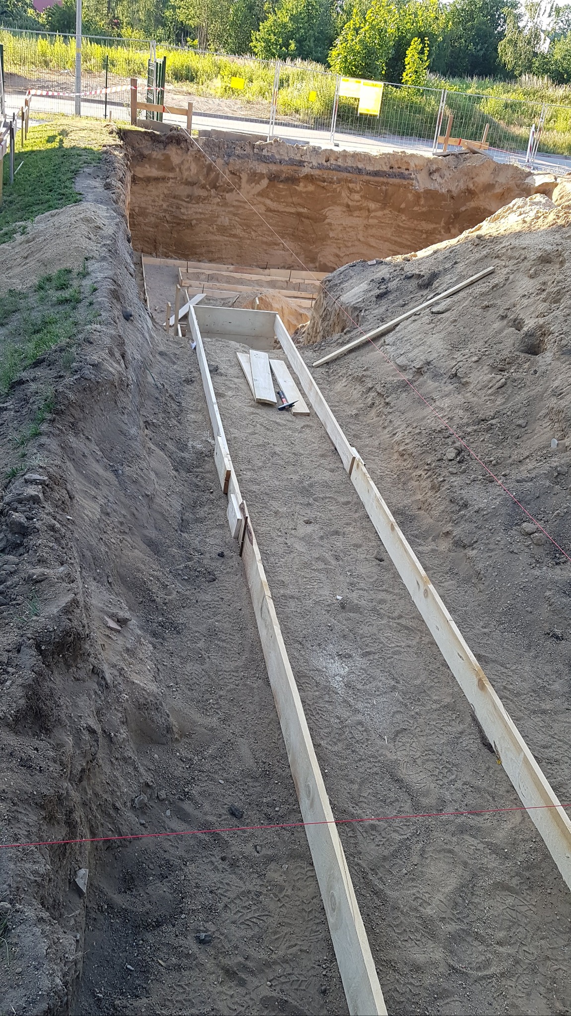 Wykop pod fundamenty z drewnianym szalunkiem, widoczny młotek i deski, ziemny brzeg wykopu z zabezpieczeniami.
