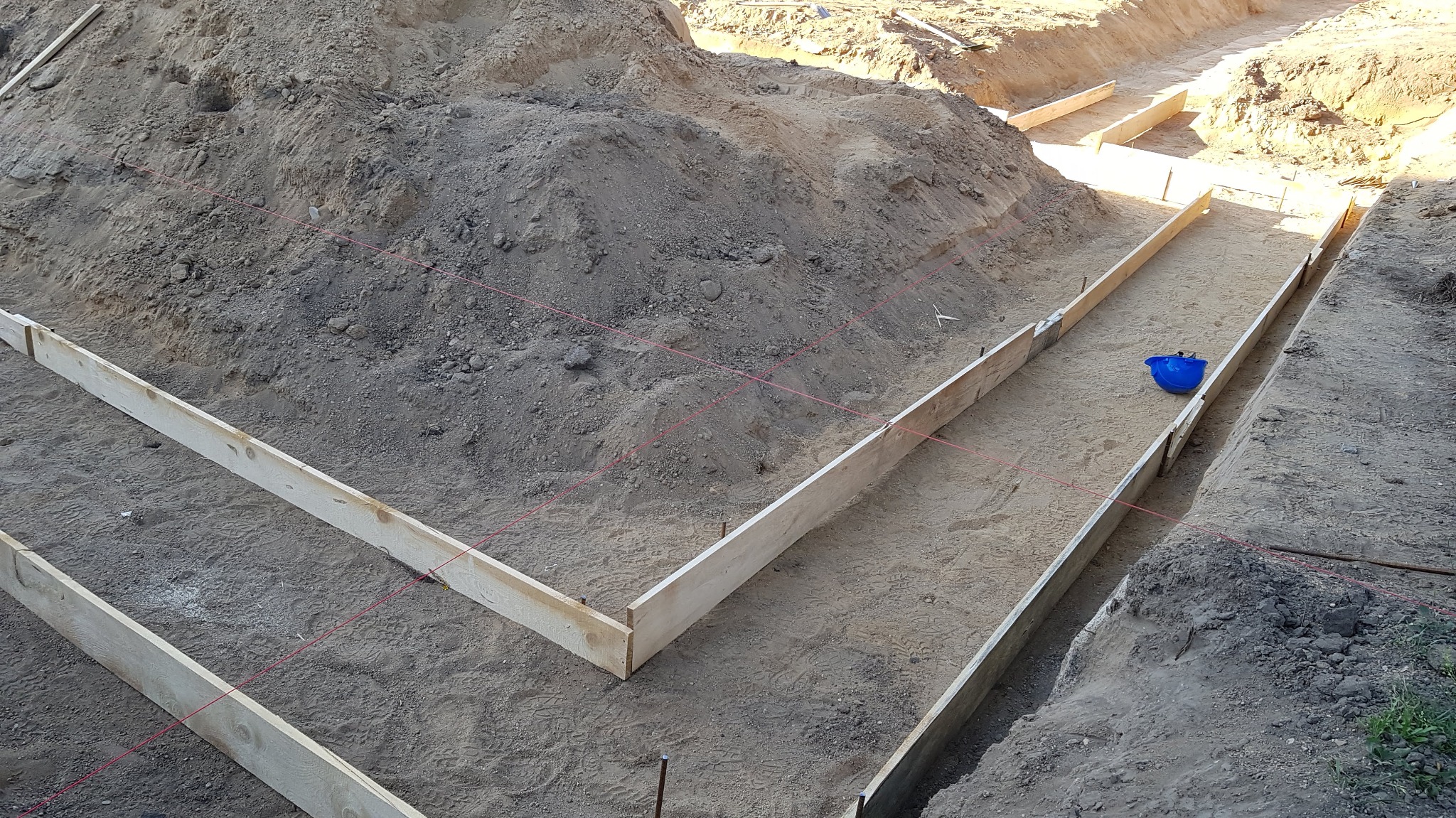 Przygotowanie wykopu i szalunku pod fundamenty domu, widoczne drewniane deski tworzące formę, wykopana ziemia i niebieskie wiadro wewnątrz szalunku, czerwone sznurki wyznaczające osie.